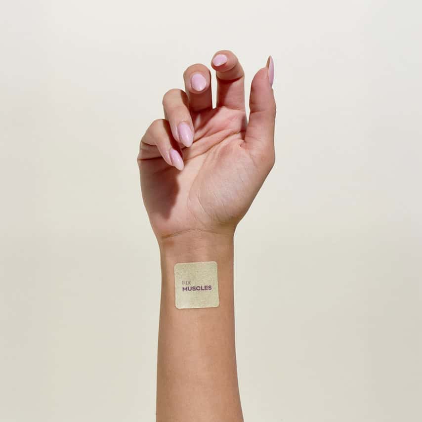 יד עם מדבקה ריבועית בצבע בז' עליה כתוב "FIX MUSCLES" מודבקת על פרק כף היד.