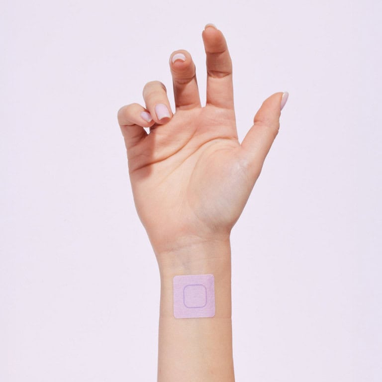 יד עם מדבקה ריבועית סגולה-בהירה על פרק כף היד, מונחת על רקע לבן.