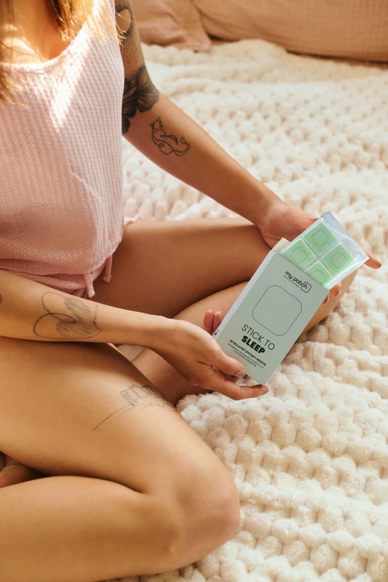 אישה יושבת על מיטה ומחזיקה בידיה בקבוק קרם עם הכיתוב "STICK TO SLEEP".